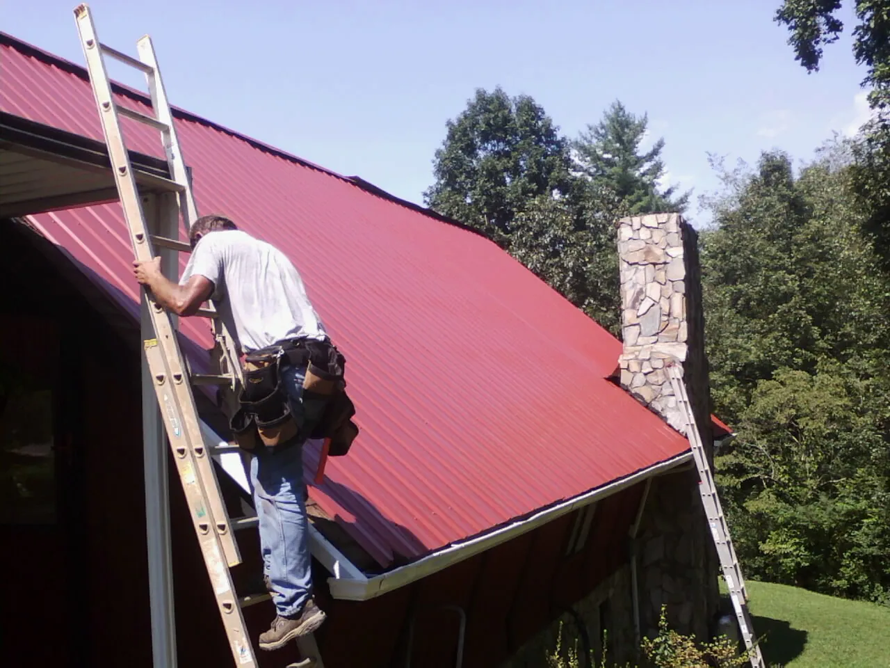 Roofer on a ladder beside a red metal roof and stone chimney — Soffit & Fascia Repair by Roofing Brothers in Sidney Center